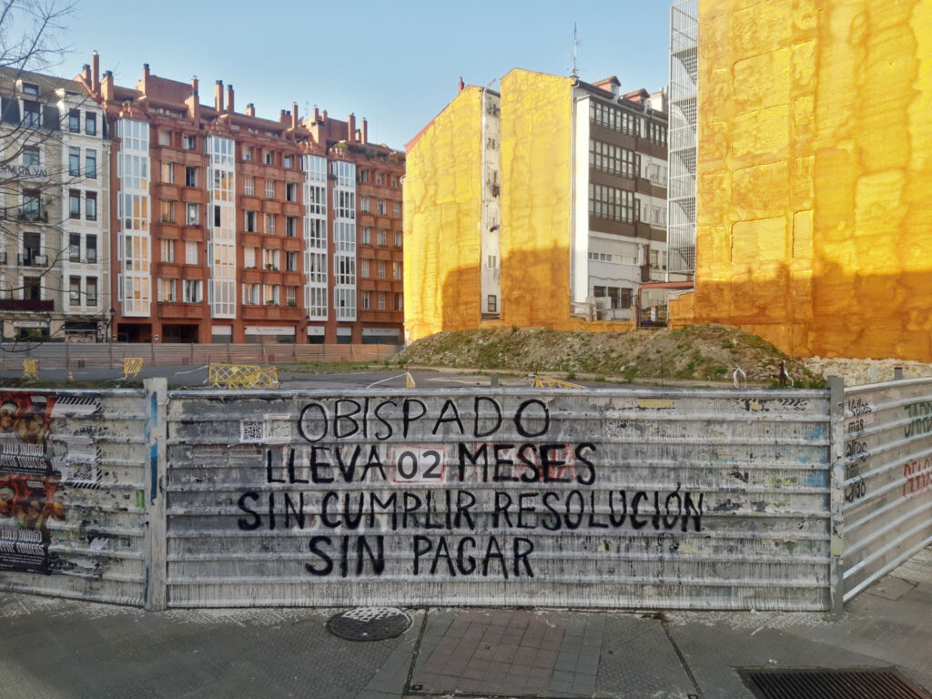 Valla de obra en la que aparece una pintada en la que se indica que el Obispado de Bilbao lleva 2 meses sin cumplir conlas resoluciones y sin pagar las multas