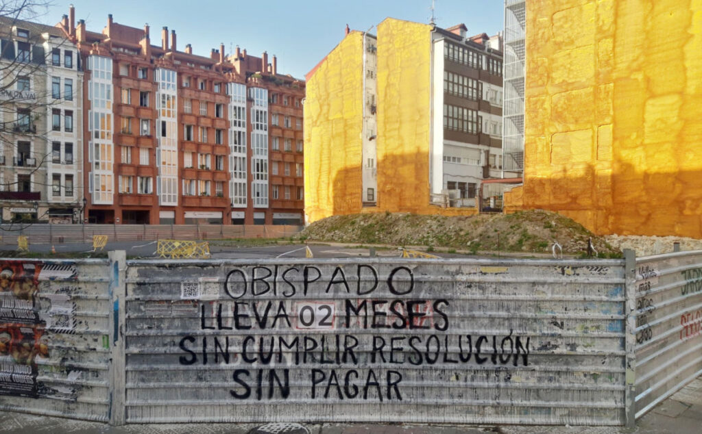 Valla de obra en la que aparece una pintada en la que se indica que el Obispado de Bilbao lleva 2 meses sin cumplir conlas resoluciones y sin pagar las multas